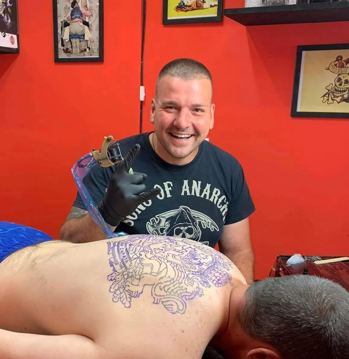 Tätowierer lächelt für die Kamera, im Hintergrund ein Tattoo in Arbeit auf dem Rücken